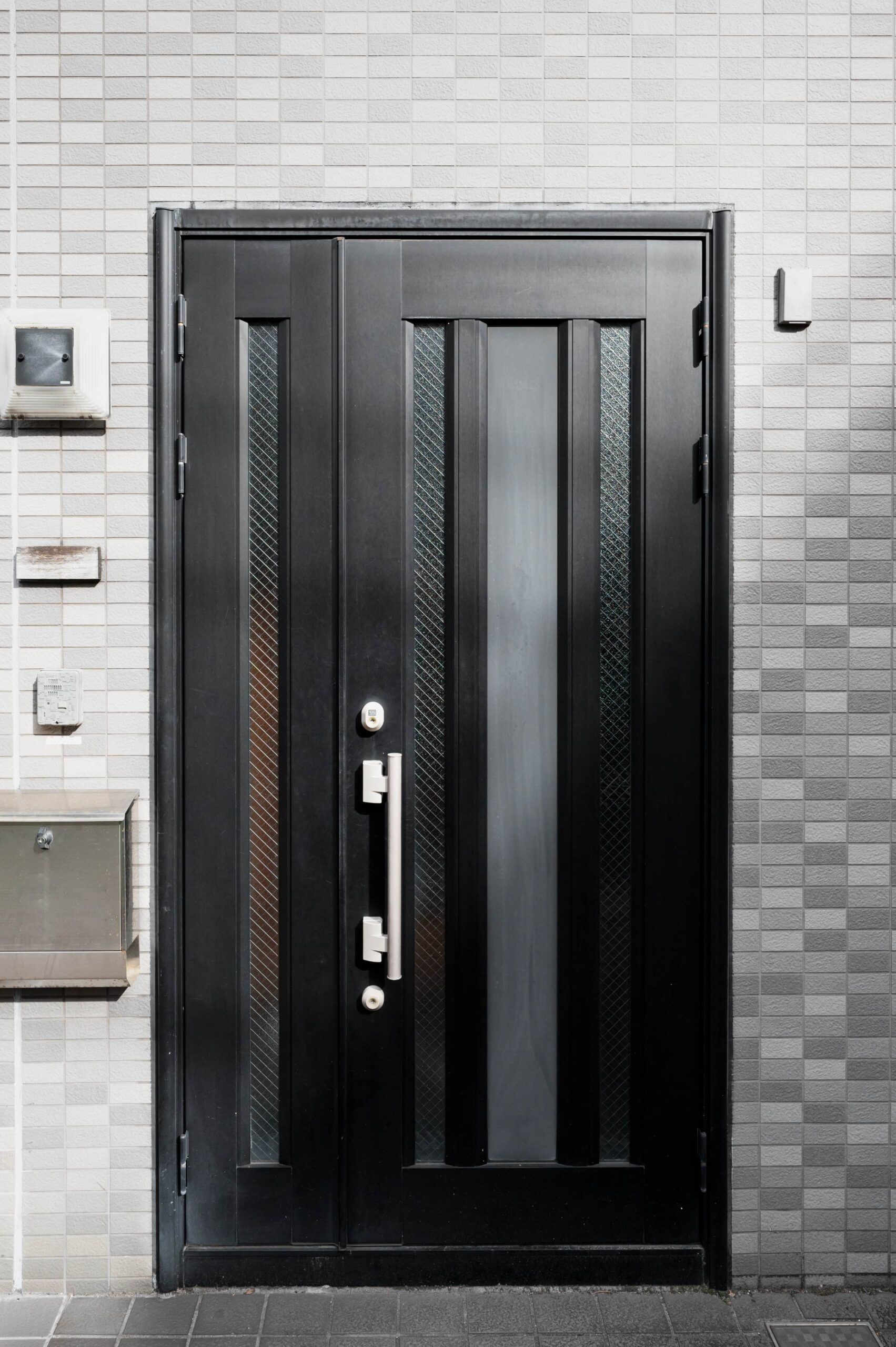 Black front door with narrow vertical glass panels and a silver pull handle, set in a white tiled wall.