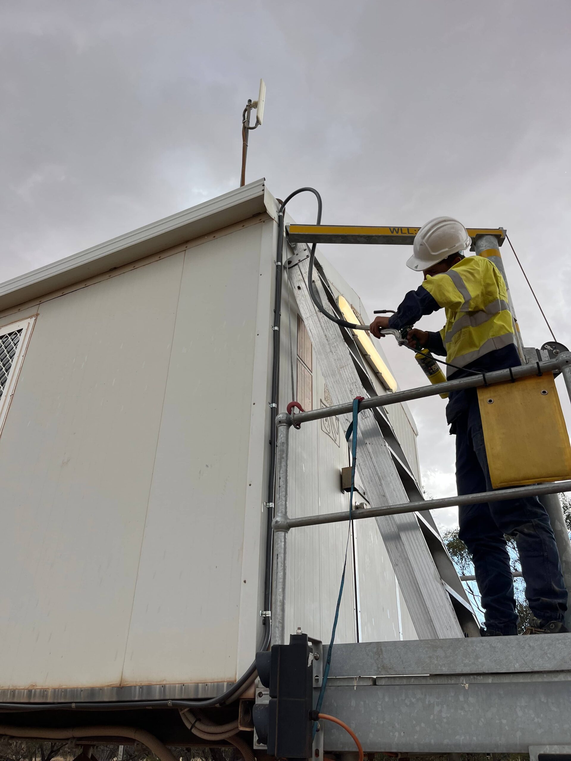 Worker installs business security components on building exterior with a power drill, wearing safety gear.