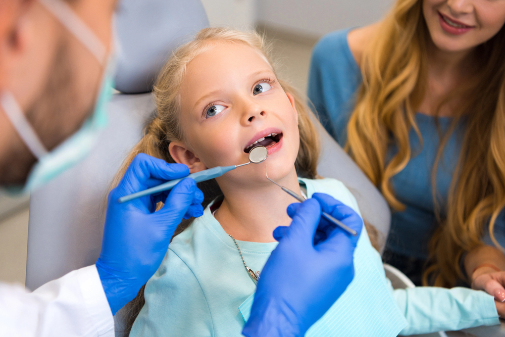 Child receiving a dental check-up from a dentist while her mother sits nearby
