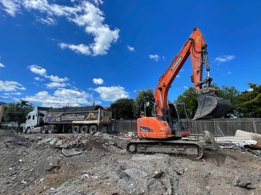 An orange Doosan excavator and a Site Demolition dump truck working on a construction site under a blue sky.