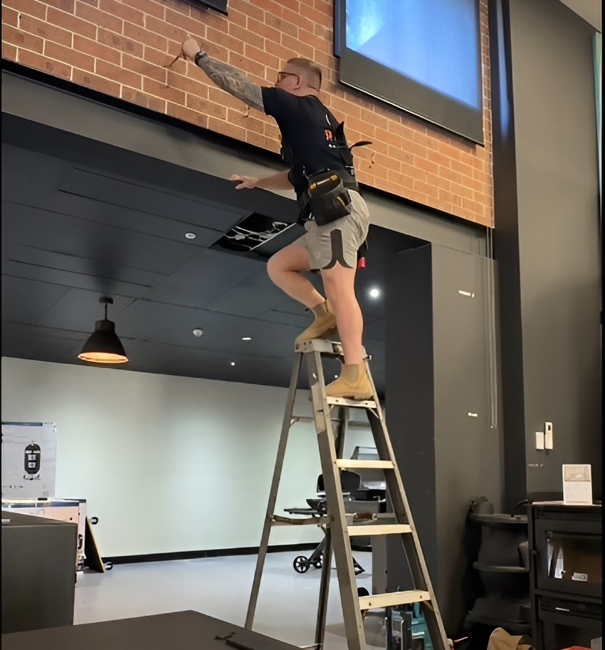 A local bucks electrician on a ladder adjusts wiring on a brick wall in a modern industrial in sydney .