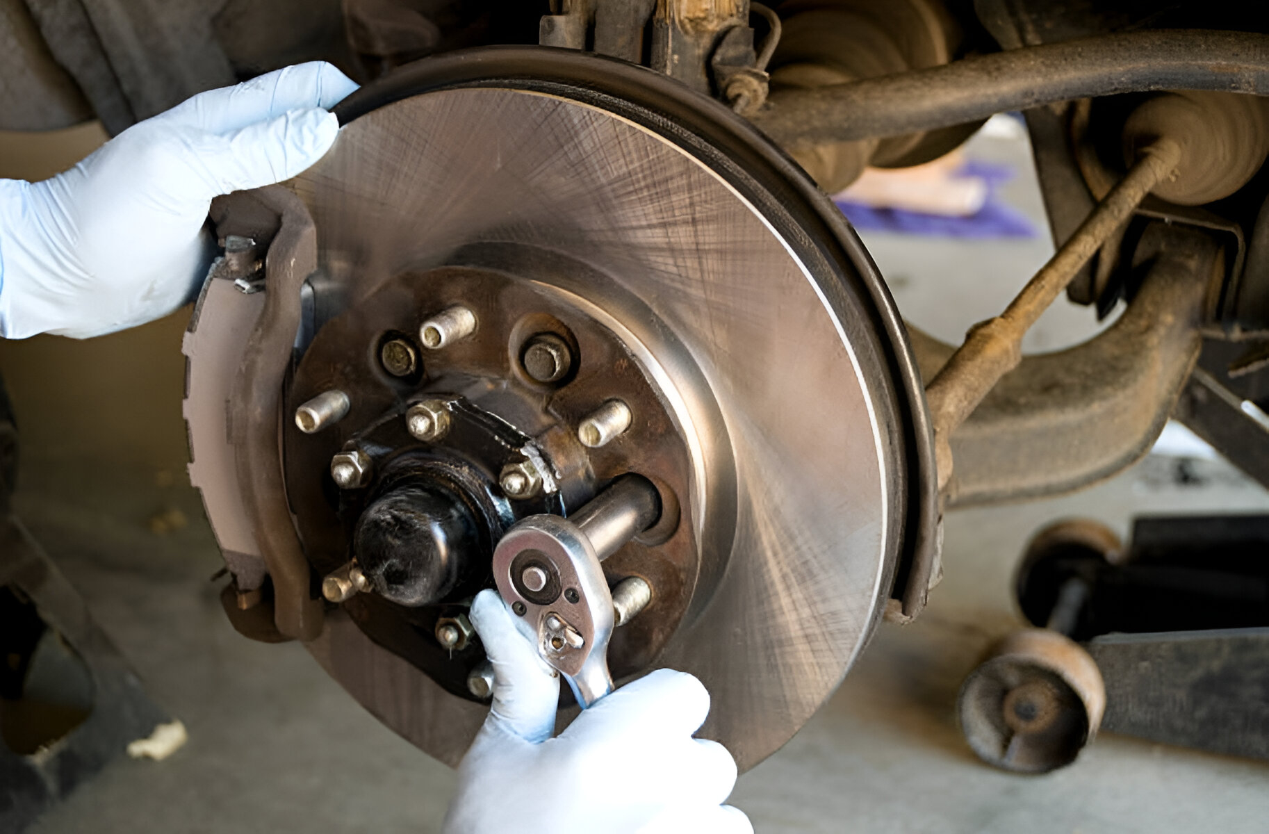A mechanic in blue gloves uses a metal ratchet to tighten a bolt on a car brake rotor during a garage brake check.