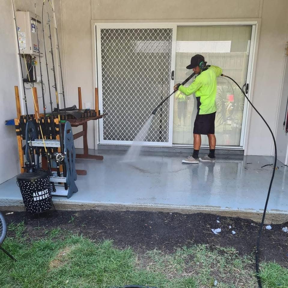 A QQCPM worker uses a machine to pressure clean a concrete patio near a glass door.