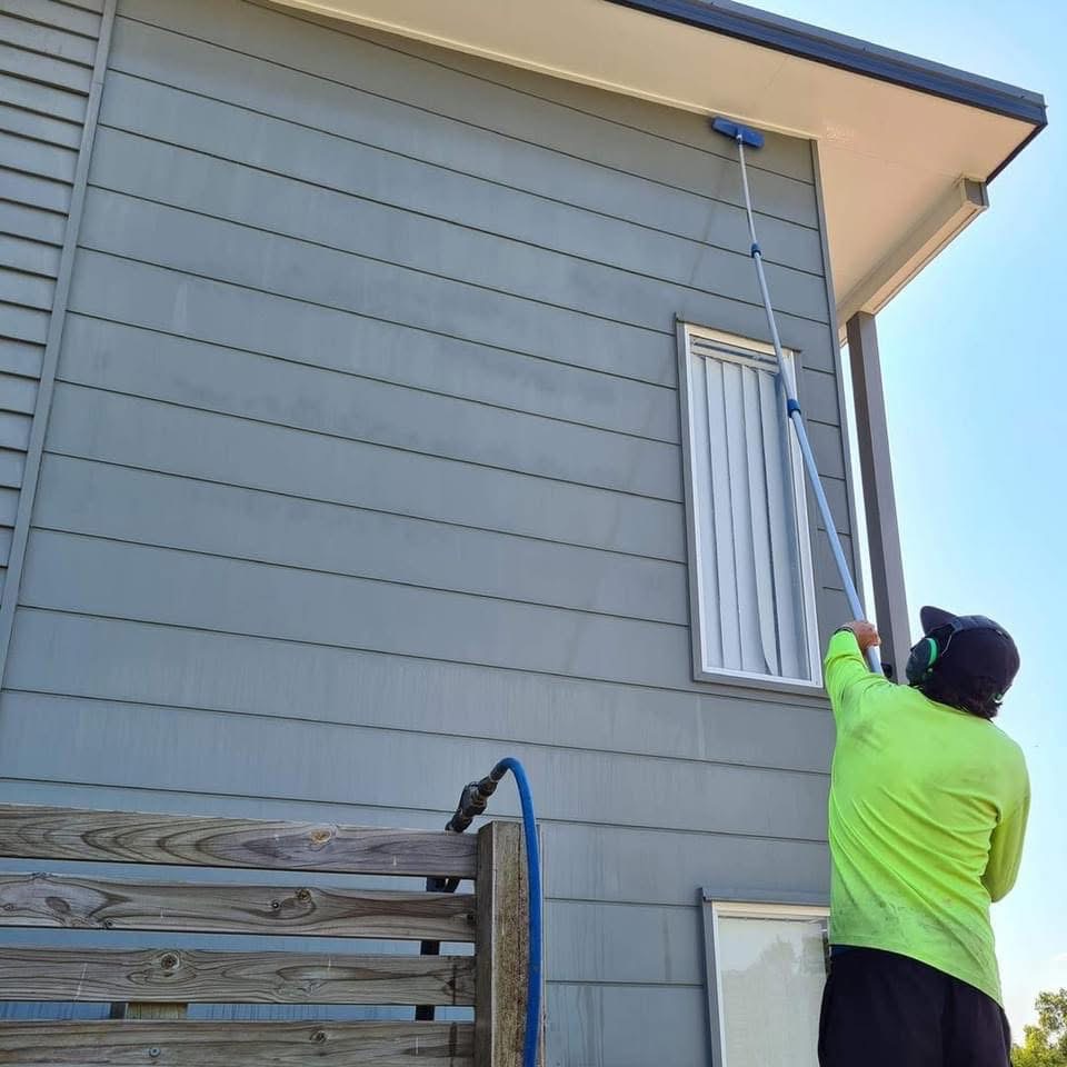 A QQCPM worker uses an extended pole brush to scrub the gray exterior siding of a two storey Brisbane home.