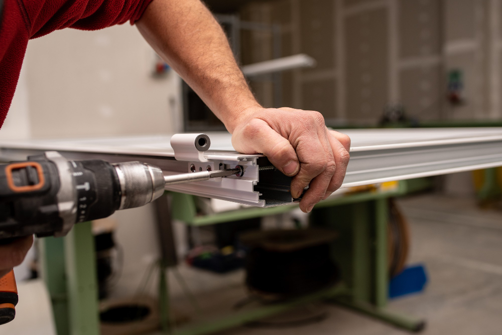Technician using a cordless drill to fasten a screw into an aluminium door frame in a workshop.