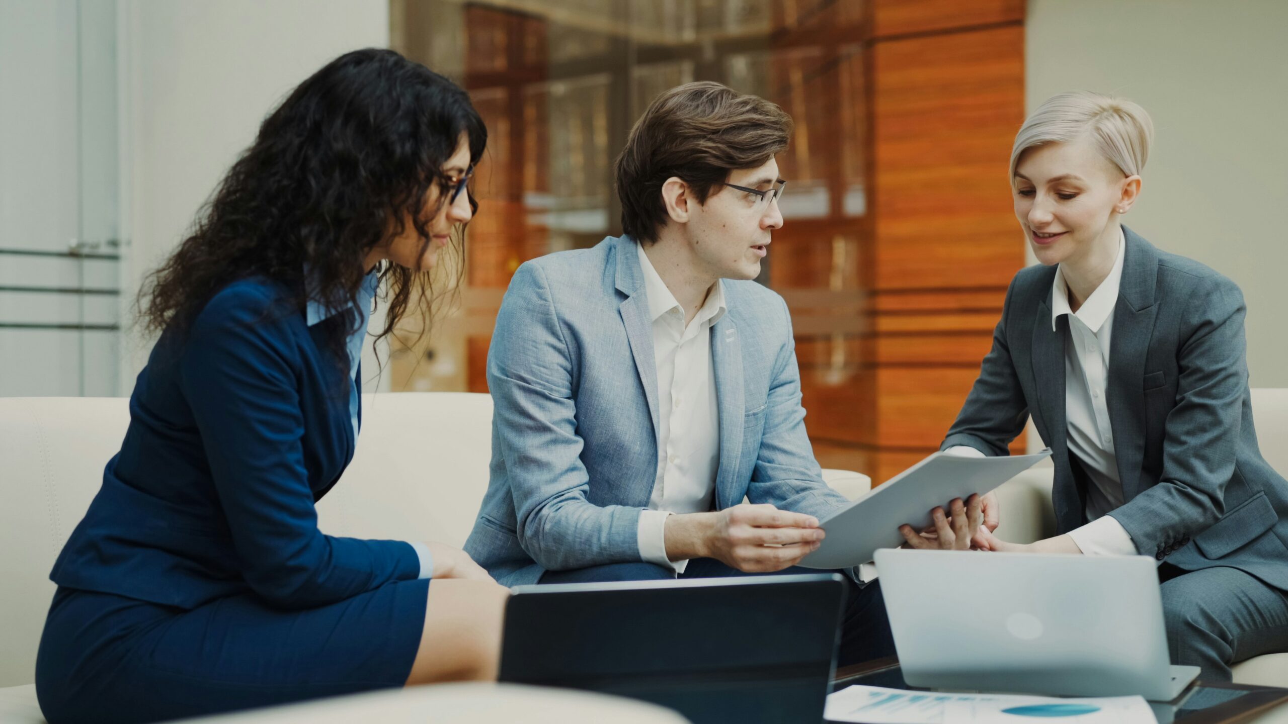 Three business professionals sitting on a sofa in an office reviewing documents during a finance business meeting.
