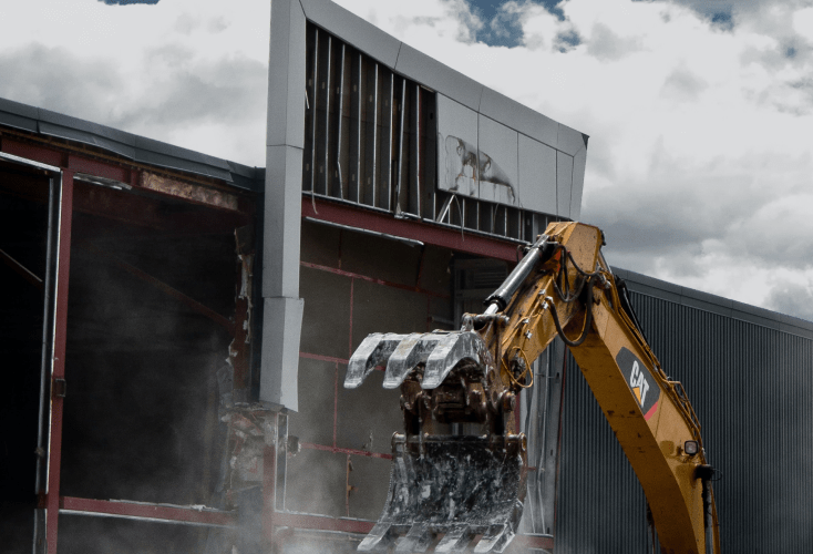 A yellow mechanical excavator performs Site Demolition on a commercial structure under a cloudy sky.